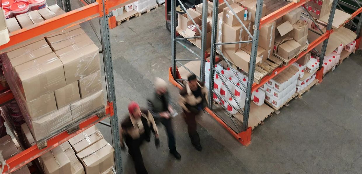 Three warehouse workers walking quickly between shelves filled with boxes and containers.