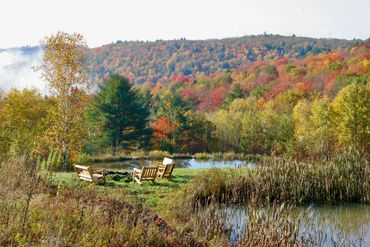 Adirondack chairs by pond  and fire pit with fall foliage at a secluded Catskills retreat