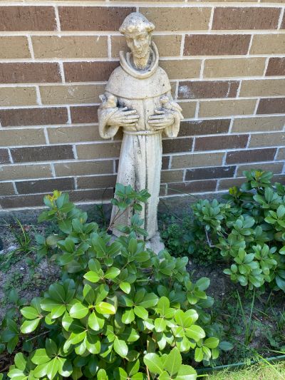 Stone statue of a monk holding birds in a garden in front of a brick wall.