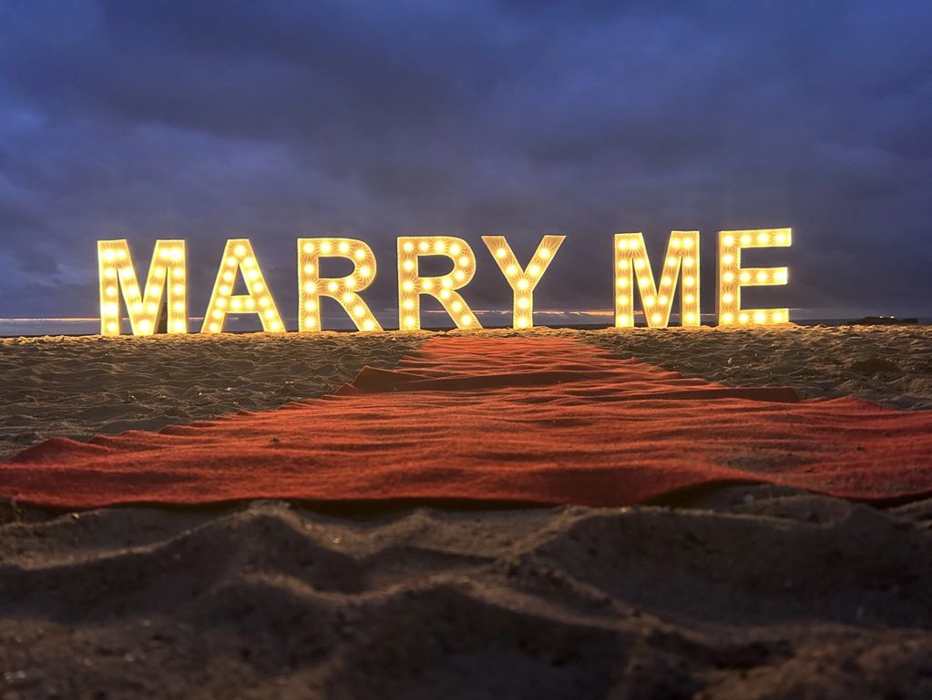 Illuminated 'MARRY ME' sign on the beach at dusk.