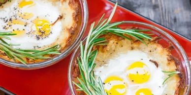 Baked eggs in glass bowls garnished with fresh rosemary on a red tray.