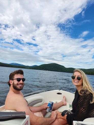 A smiling couple enjoys a boat ride on a lake surrounded by green hills and a partly cloudy sky.