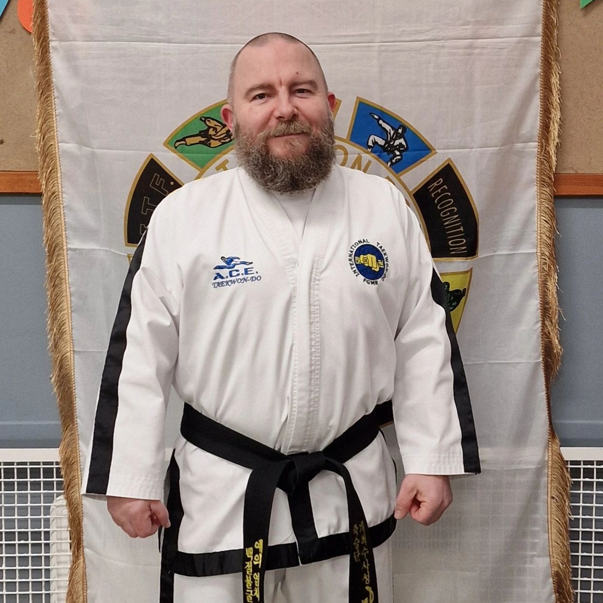 Bearded man in Taekwondo uniform with black belt stands confidently indoors.