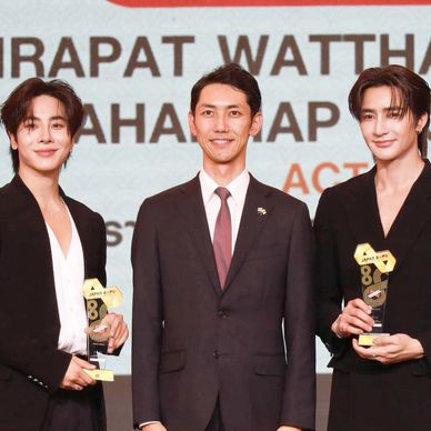 Two men holding trophies stand beside a suited man at an award event.
