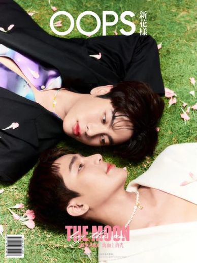 Two young men lying on grass surrounded by pink petals, gazing at each other.