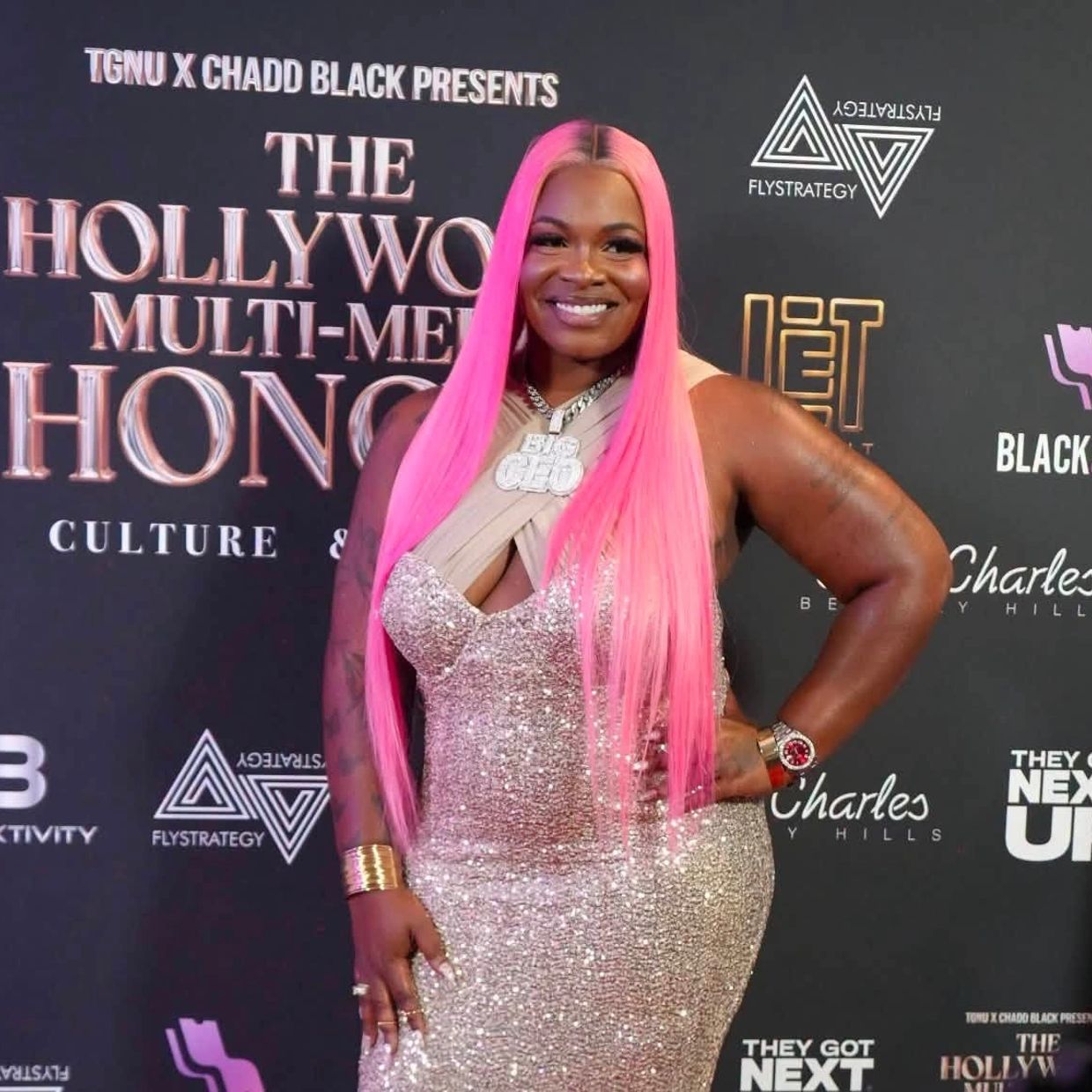 Woman with long pink hair in a sparkling dress smiles on red carpet.