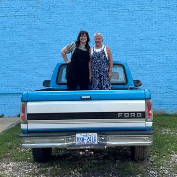 Two women stand together in the bed of a blue, vintage Ford truck in front of Altamont's blue wall. 