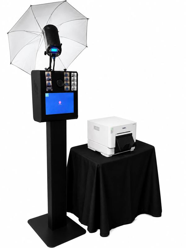 Photo booth setup with camera, umbrella light, touchscreen, and printer on black stand and table.