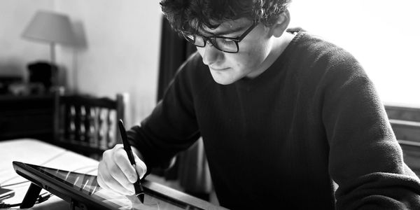 Young man drawing on a digital tablet at a desk by a window.