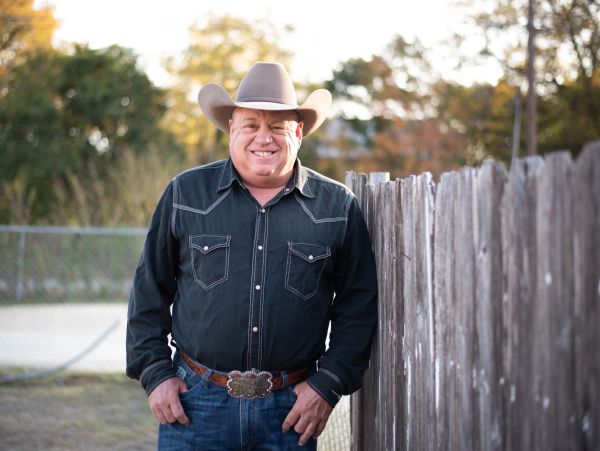 Man wearing a cowboy hat and western shirt by a wooden fence.