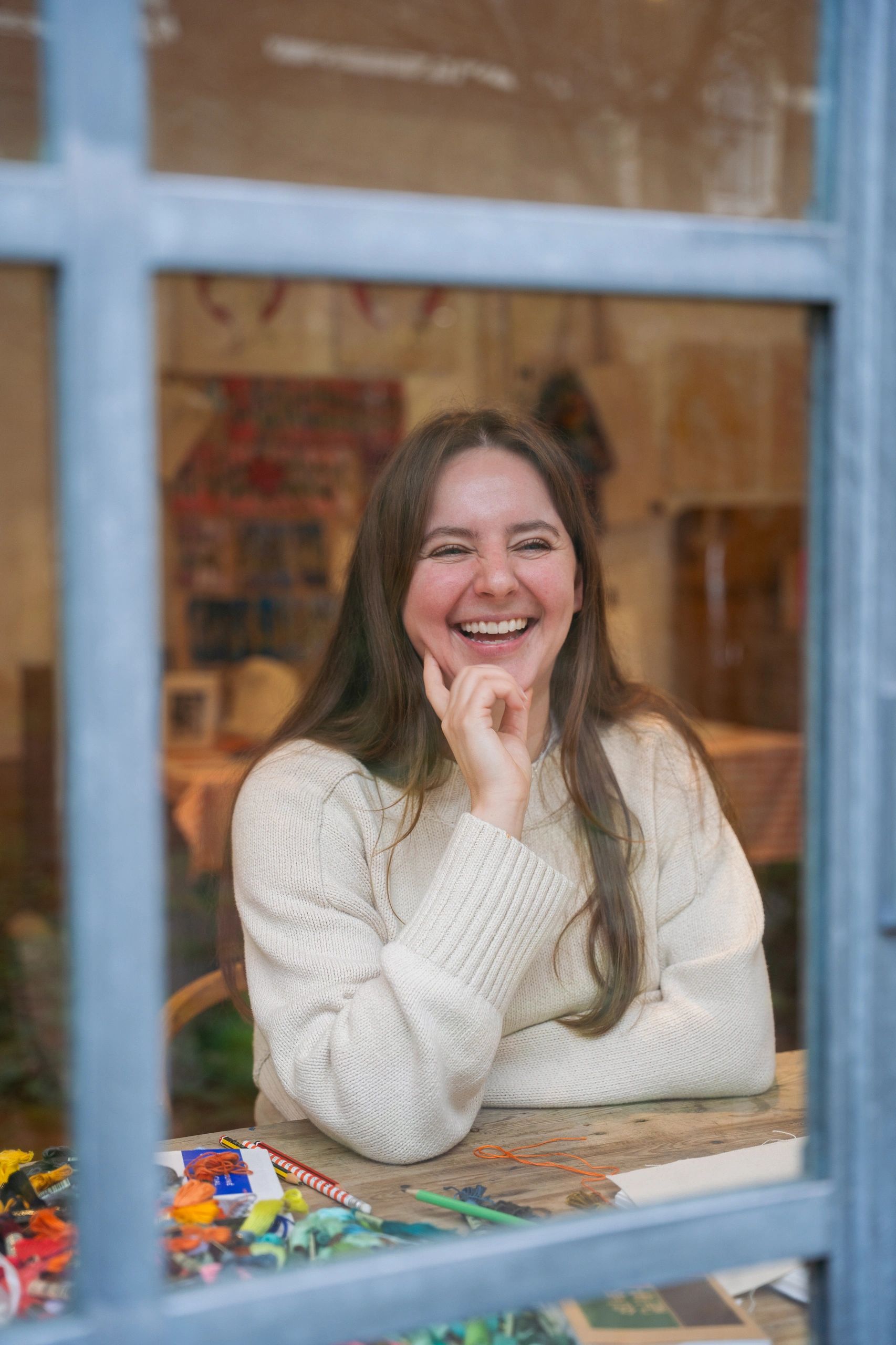 A woman smiling behind a window at a table with art supplies.