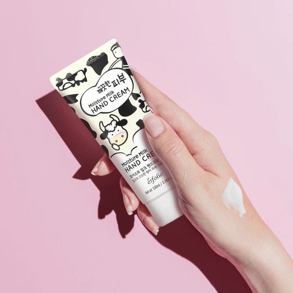 Hand holding a cute cow-patterned Moisture Milk Hand Cream tube on pink background.