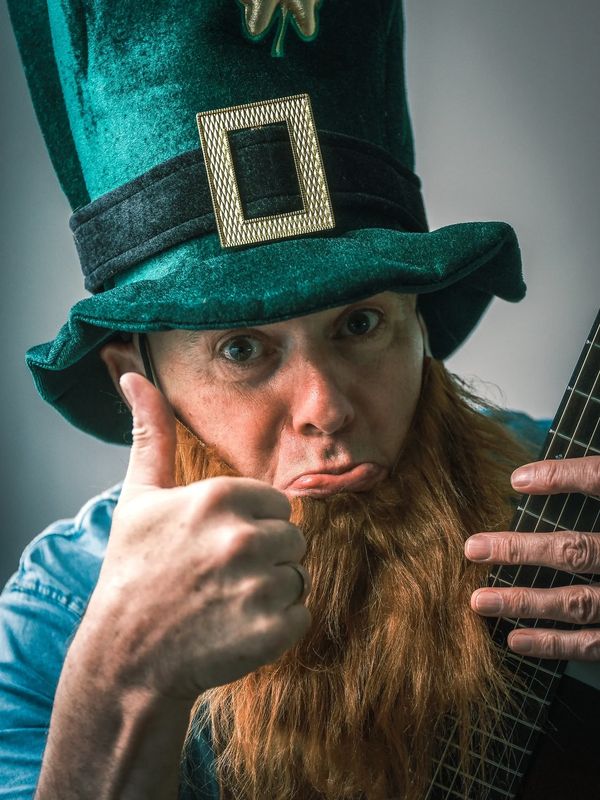 Man in a leprechaun hat giving thumbs up and holding a guitar.