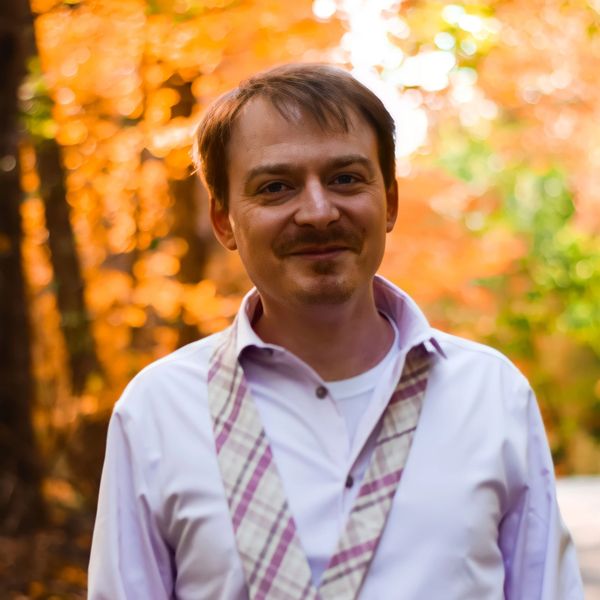 A man in a white shirt and loosened tie stands smiling outdoors in autumn.