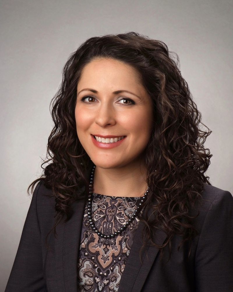 Dana Nevedal, Ph.D. Smiling brunette woman wearing black jacket, patterned blouse, pearl necklace.