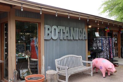 Exterior of a store named BOTANICA with a bench and a decorative pig outside.