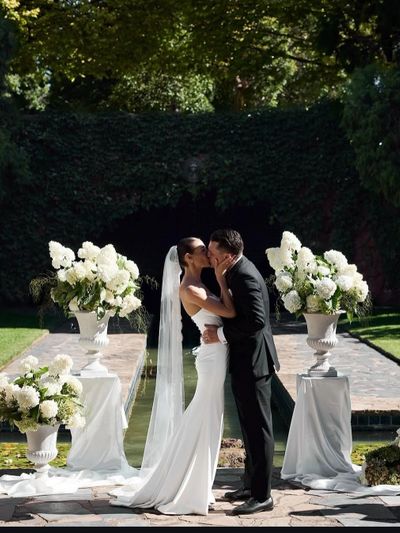 Bride and groom share a kiss during an outdoor wedding ceremony surrounded by white floral arrangements.