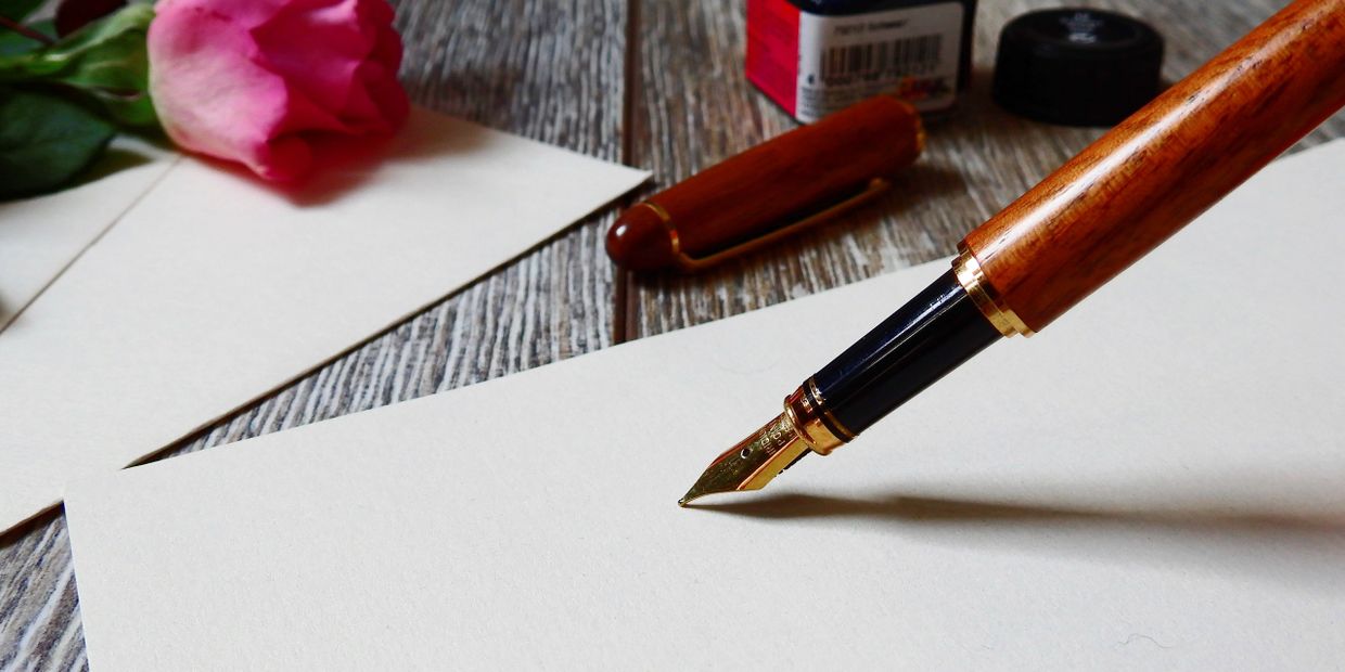 Calligraphy pen, poised on white sheet of paper, with envelope nearby, on wooden desk.