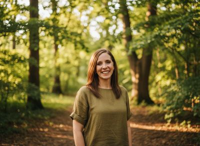 Smiling woman in green shirt standing in a sunlit forest path.