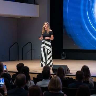 Woman speaking on stage at a conference with a large digital screen behind her.