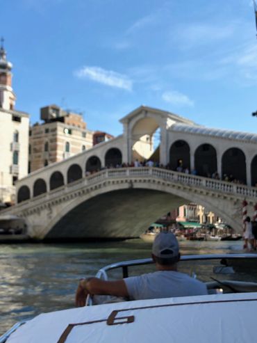 Man relaxing on a boat near a historic Venetian bridge under blue sky.