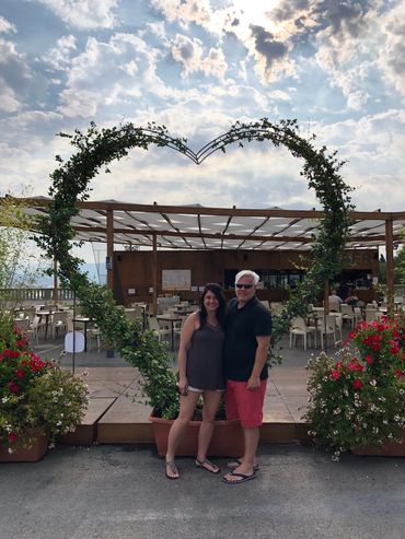 Couple posing happily inside a heart-shaped plant frame at an outdoor café.
