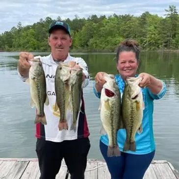Two people proudly hold large fish by a lakeside dock.