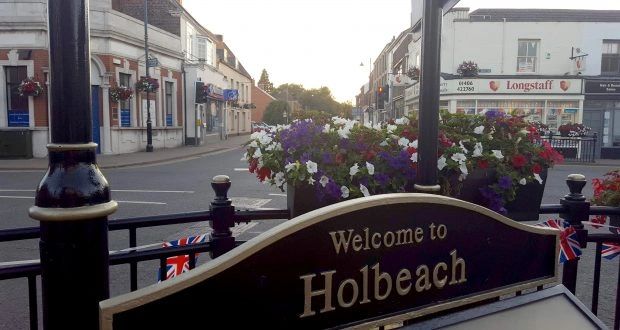 Welcome sign for Holbeach with flowers and street view in the background.