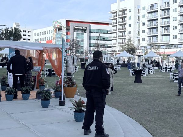Security guard monitors an outdoor event with tents and attendees in an urban area.
