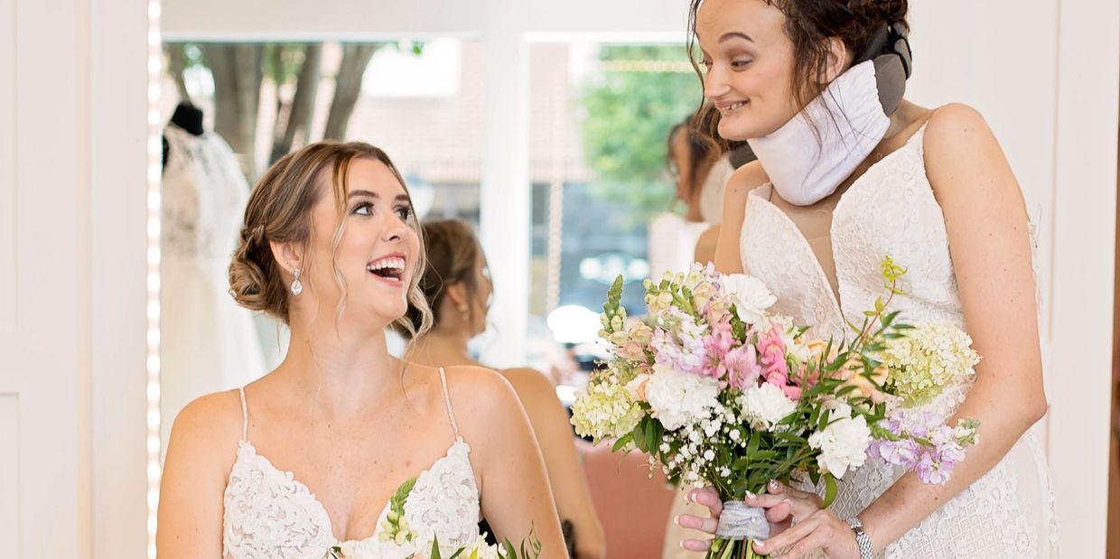 Emma and Macy, two young white women, smile at eachother and laugh. Both are wearing a wedding dress
