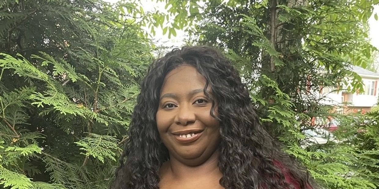 Smiling woman with long curly hair standing outdoors near green foliage.