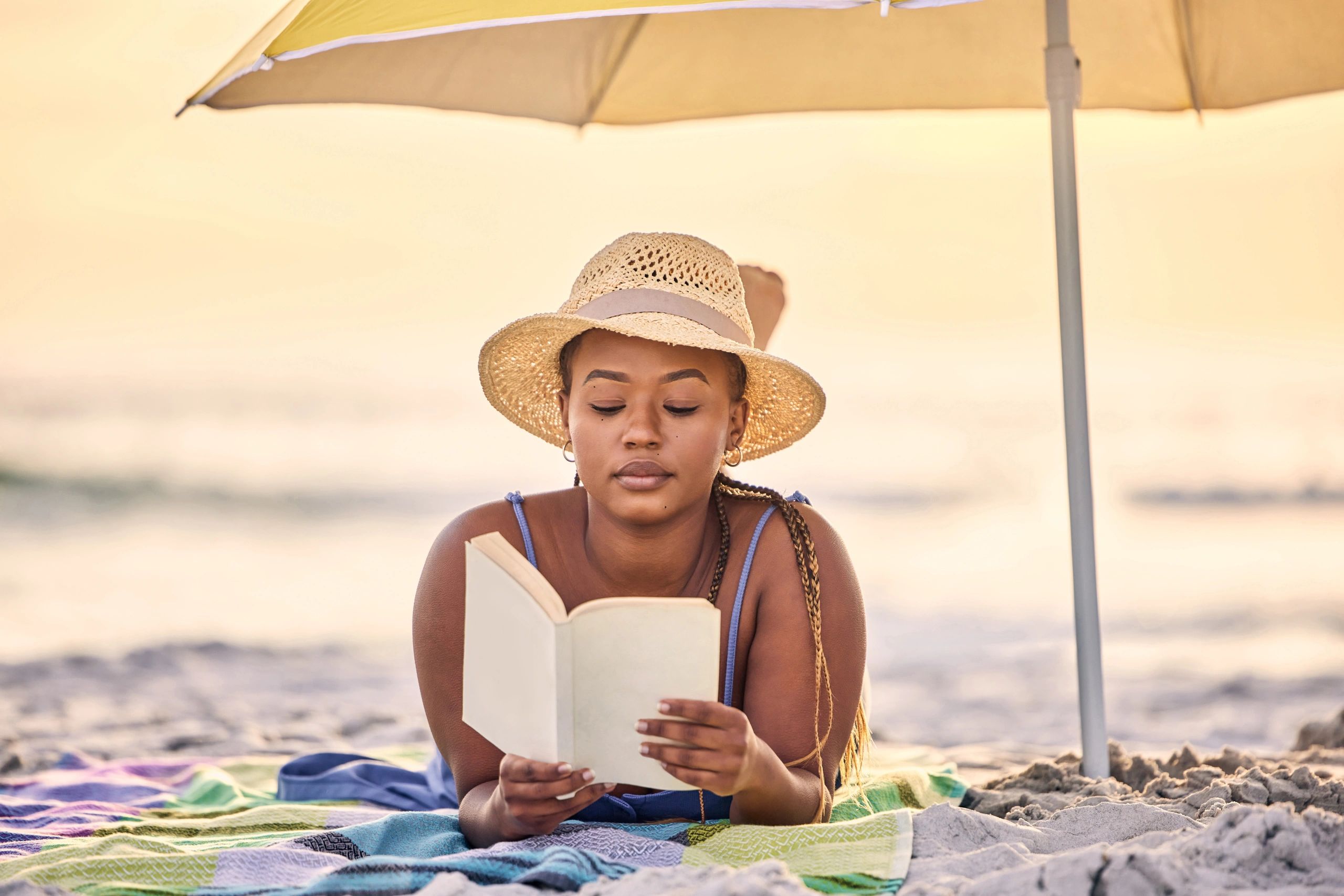 Woman reading a book under a beach umbrella on a colorful blanket.