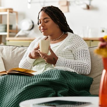 Woman relaxing on couch with a cup and a book, covered by a green blanket.