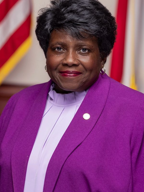 Portrait of a smiling woman in a purple blazer with flags in the background.