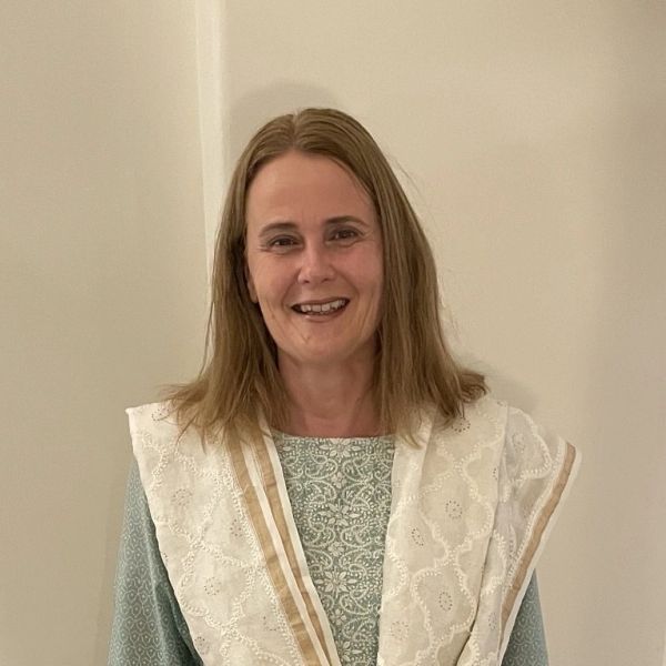 A woman with light brown hair smiling, wearing a light green top and a white shawl with gold trim.
