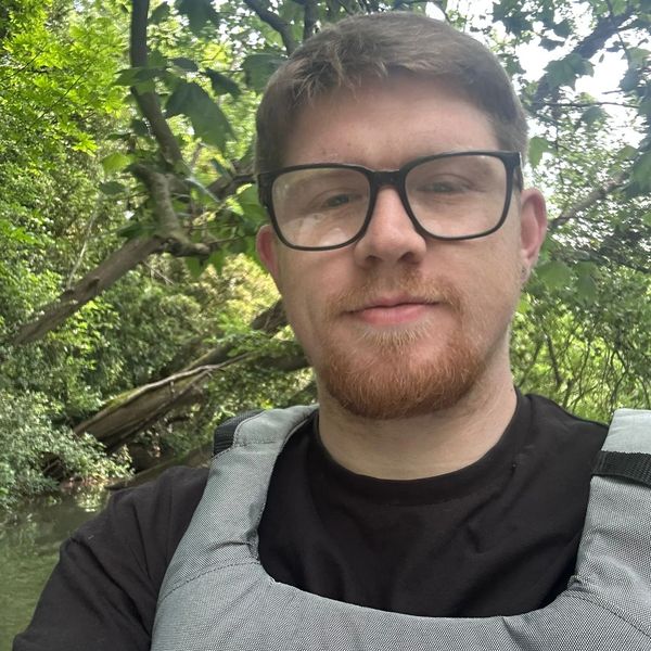 A man with glasses and a beard wearing a life jacket in a green, wooded area.