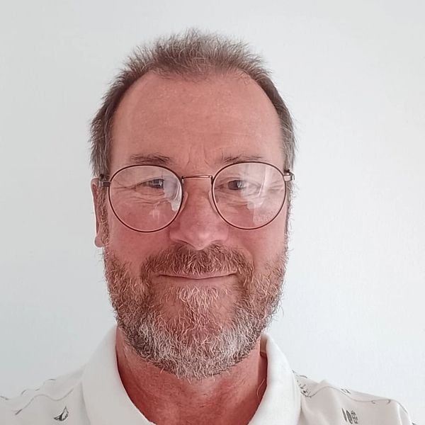 Smiling man with glasses and a patterned white shirt against a plain background.