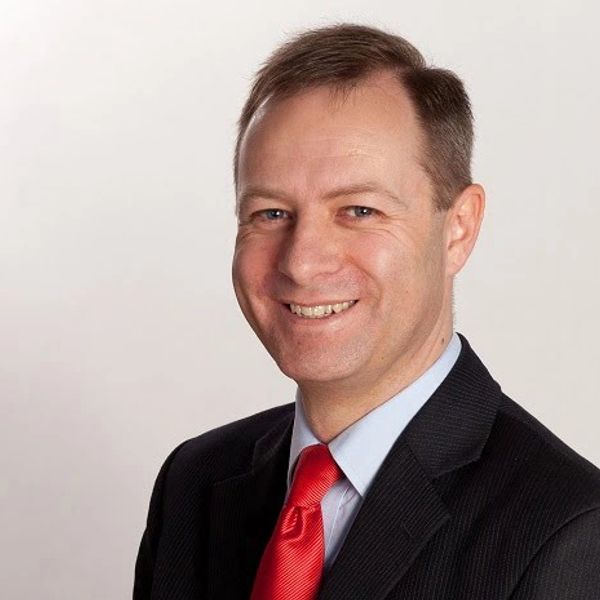 Smiling man in a suit with a red tie against a plain background.