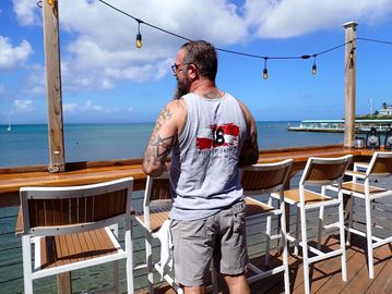 Man with tattoos standing by the ocean at a bar.