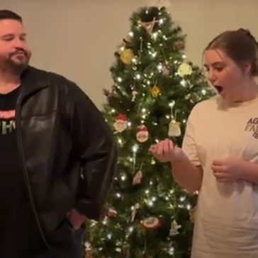A man and woman react differently near a decorated Christmas tree indoors.