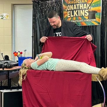 Magician Jonathan Elkins performs a levitation trick with a young girl lying on a table covered in a red cloth.
