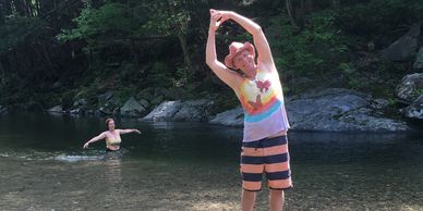 A boy in colorful clothes stretching by a river with a woman in the water behind him.