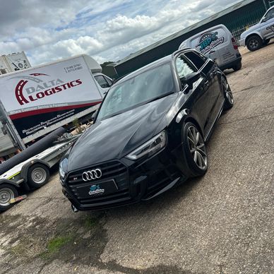 Black Audi parked on a lot with logistics trucks in background.