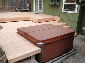 New wooden deck built around a covered hot tub beside a green house.