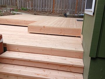 Newly built wooden deck with steps leading up to it beside a green house.
