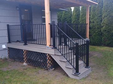 A small covered wooden porch with black railings attached to a house.