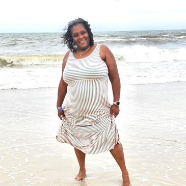 Woman smiling and standing barefoot on the beach with waves in the background.
