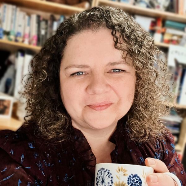 Smiling woman holding a floral-patterned mug in a cozy room with bookshelves.