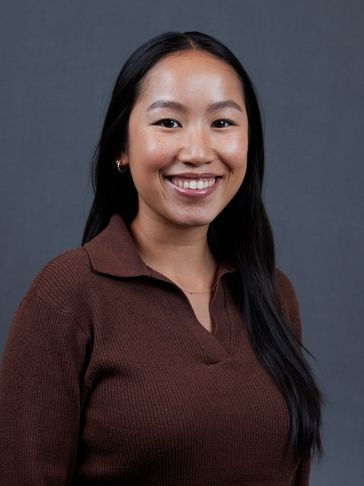 Smiling woman with long black hair and brown sweater against a gray background.