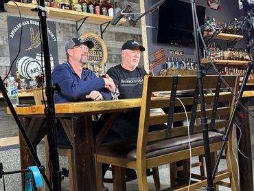 Two men sitting at a wooden bar counter with microphones and recording equipment.